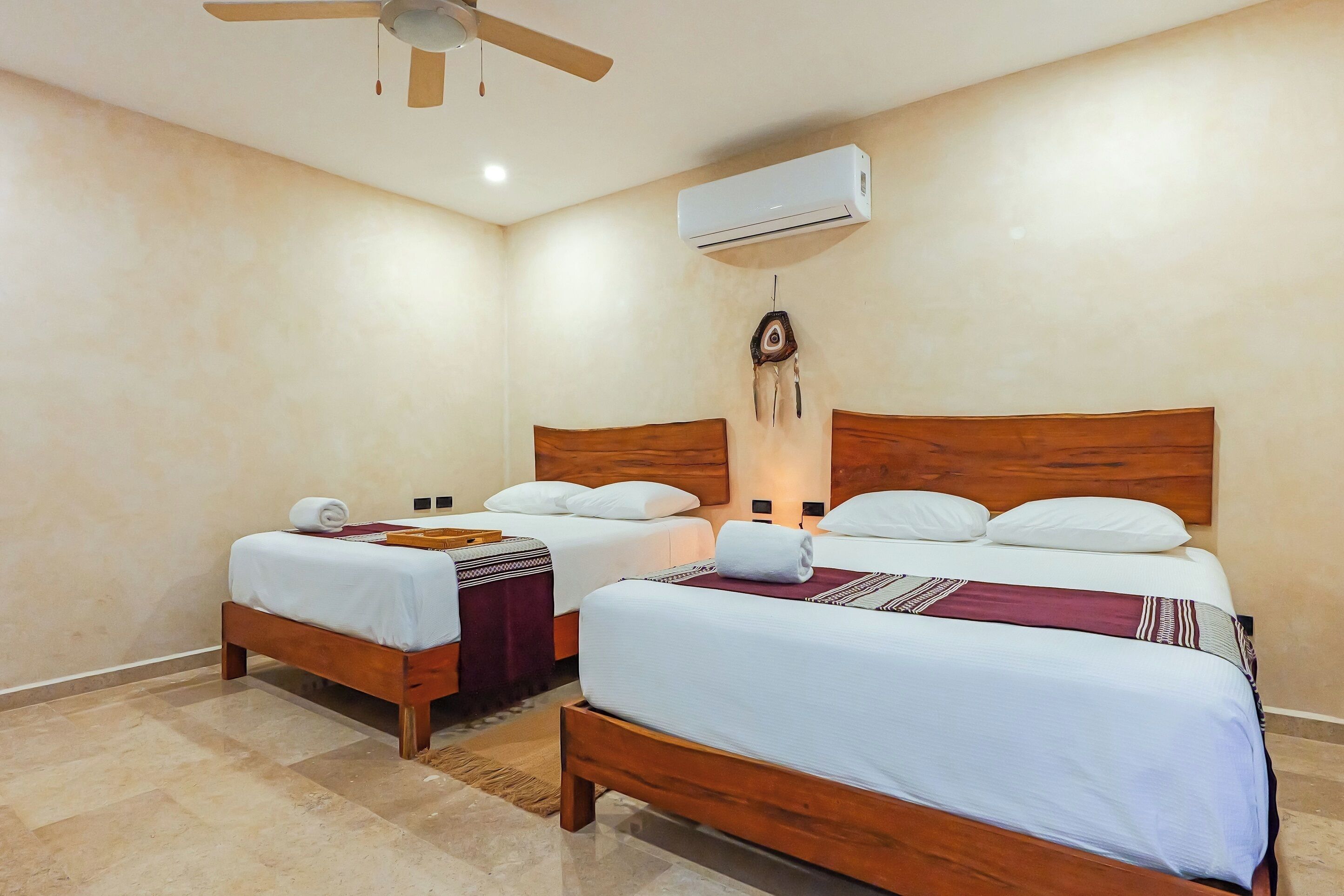 A hotel room with two wooden beds, white linens, a ceiling fan, an air conditioning unit, and a small rug.