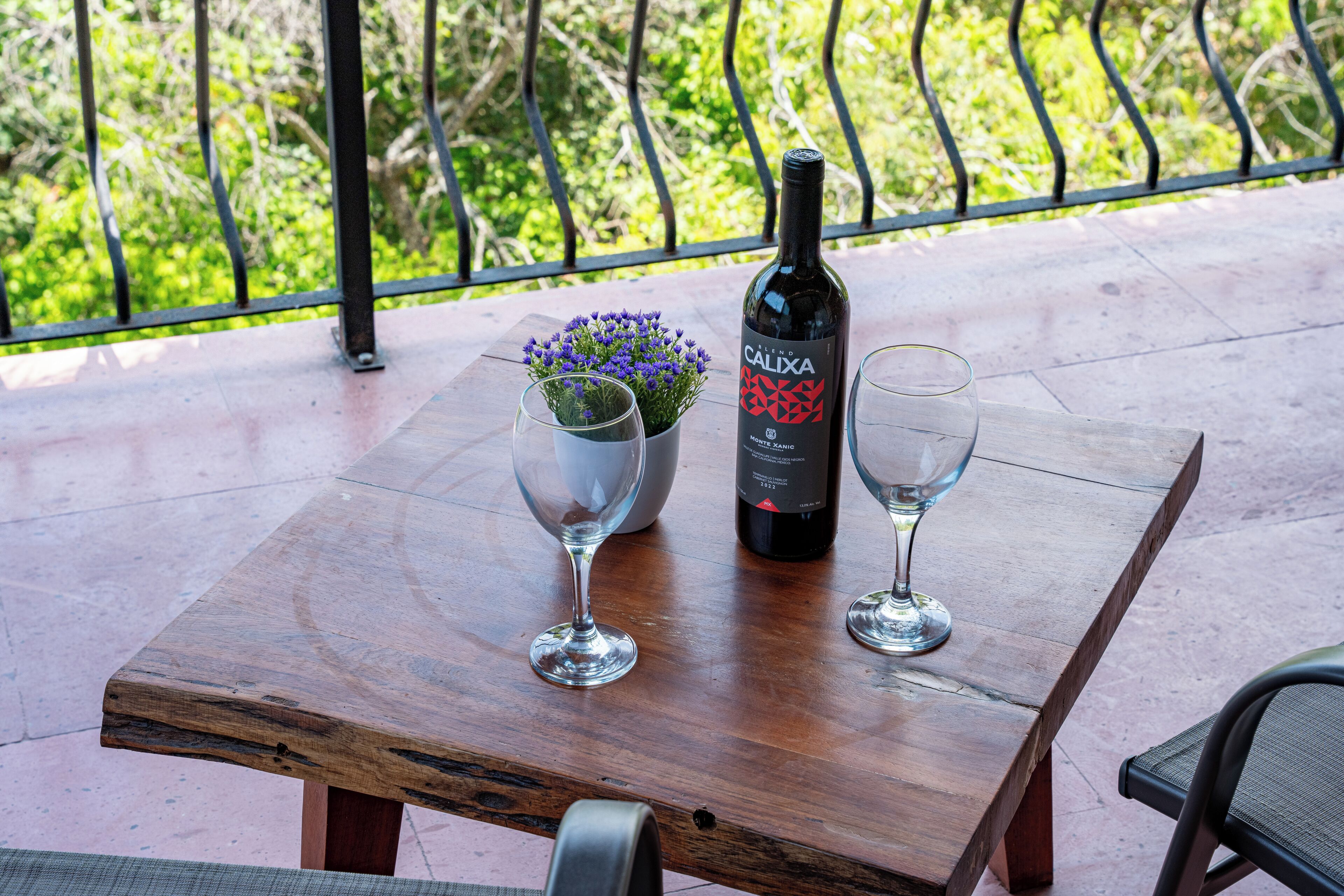 Un tavolo di legno con una bottiglia di vino e due bicchieri su un balcone con ringhiera metallica e vegetazione sullo sfondo.