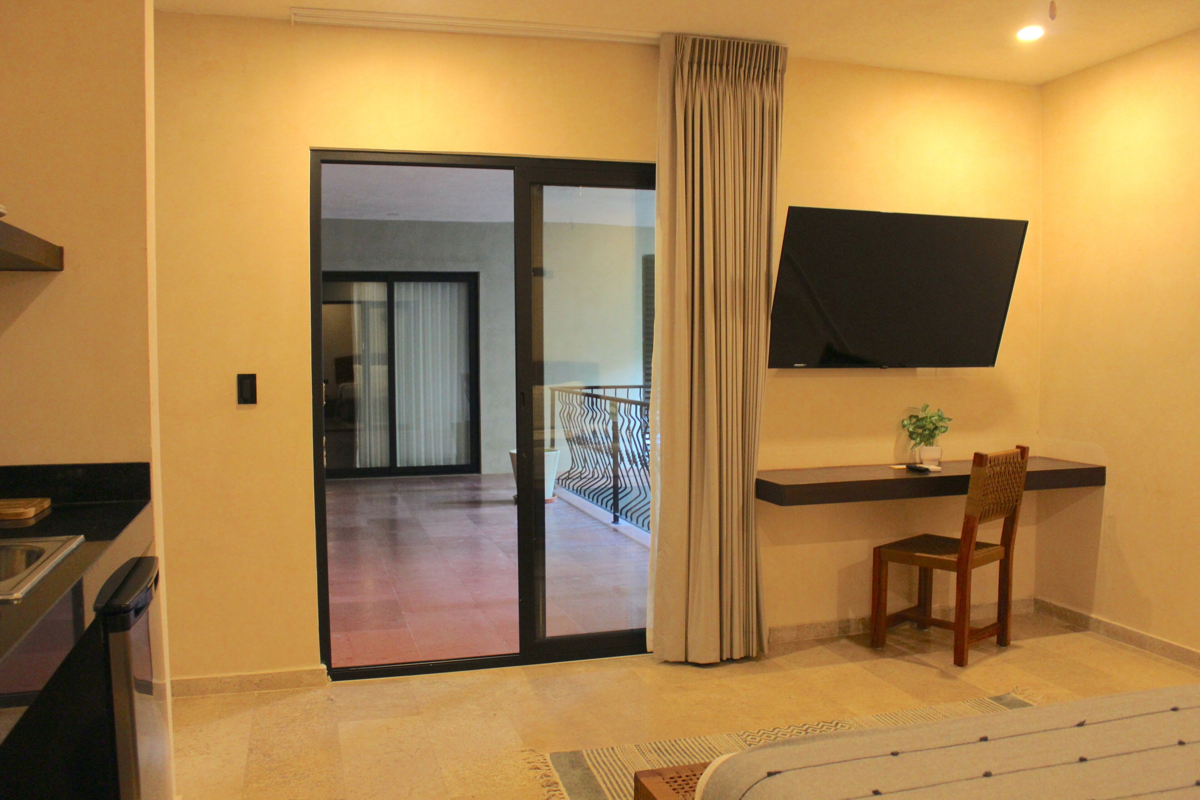 A hotel room with a sliding glass door leading to a balcony, a flat-screen TV mounted on the wall, a wooden chair, and a small table with a plant.