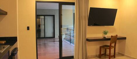A hotel room with a sliding glass door leading to a balcony, a flat-screen TV mounted on the wall, a wooden chair, and a small table with a plant.