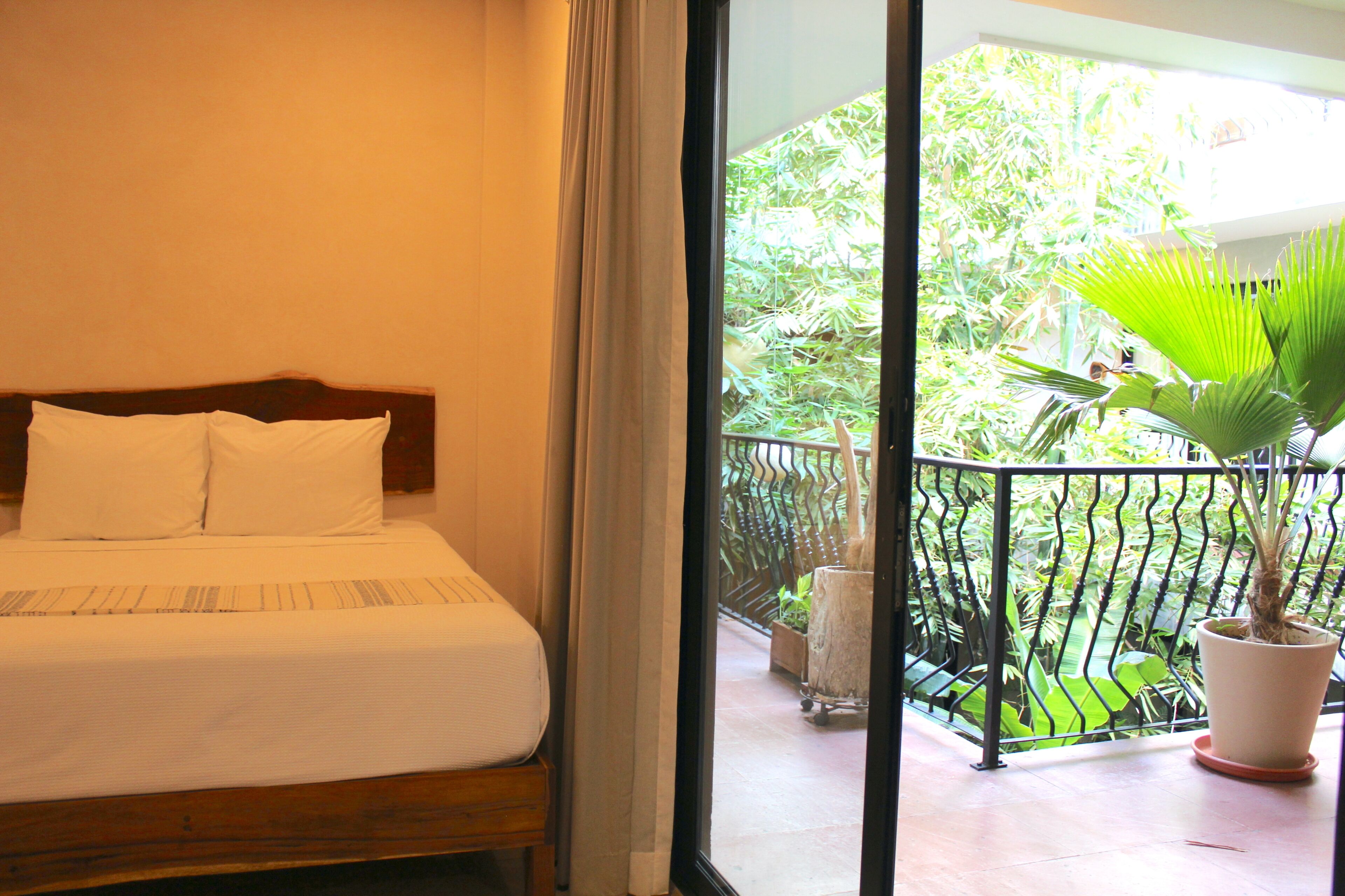 A hotel room with a bed, a potted plant, and a view of greenery through a balcony door.