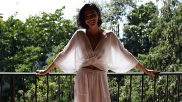 A person standing on a balcony with a railing, wearing a white dress, with greenery in the background.