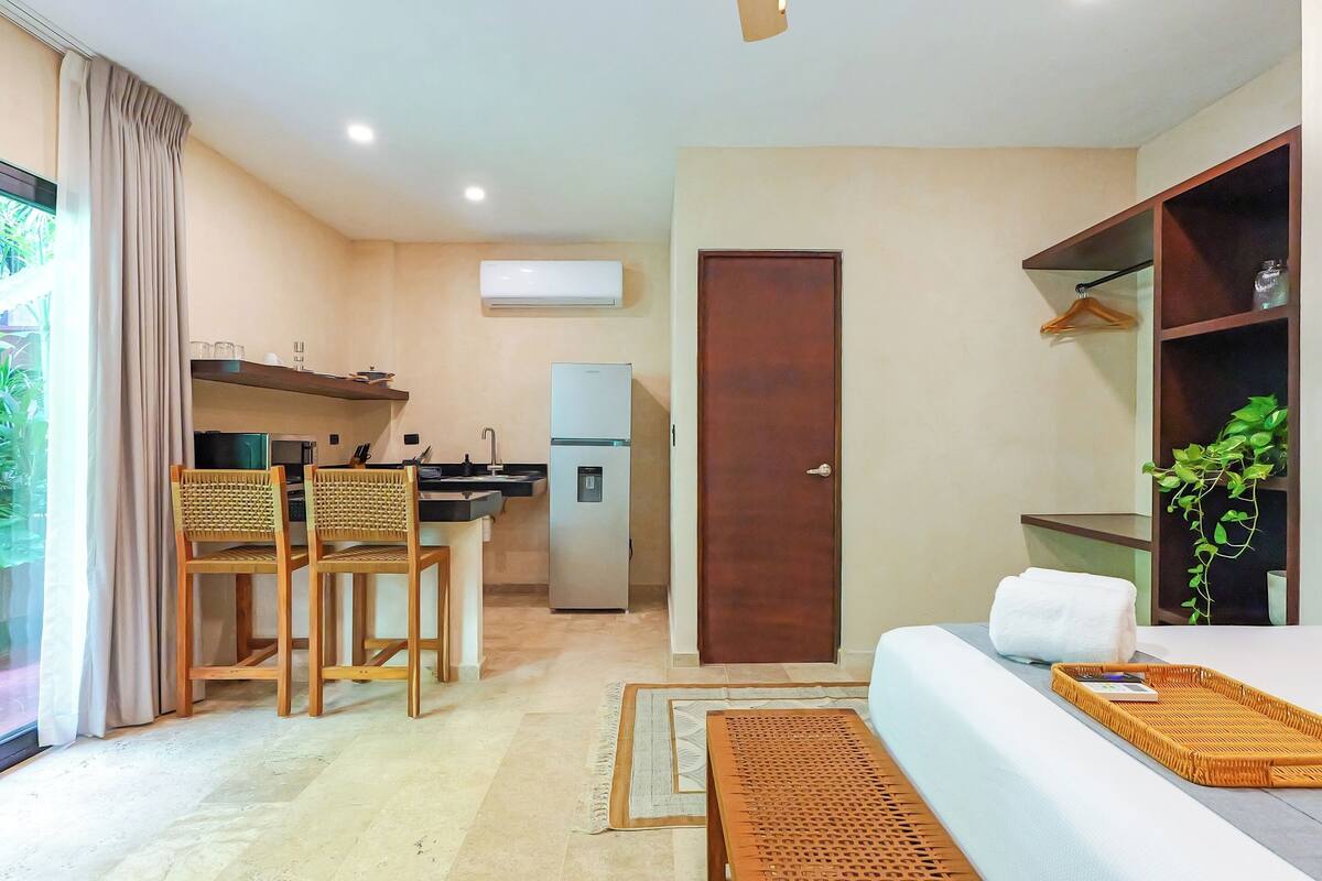 A modern hotel room with a kitchenette, a dining area with chairs, a bed with a tray, and a door.