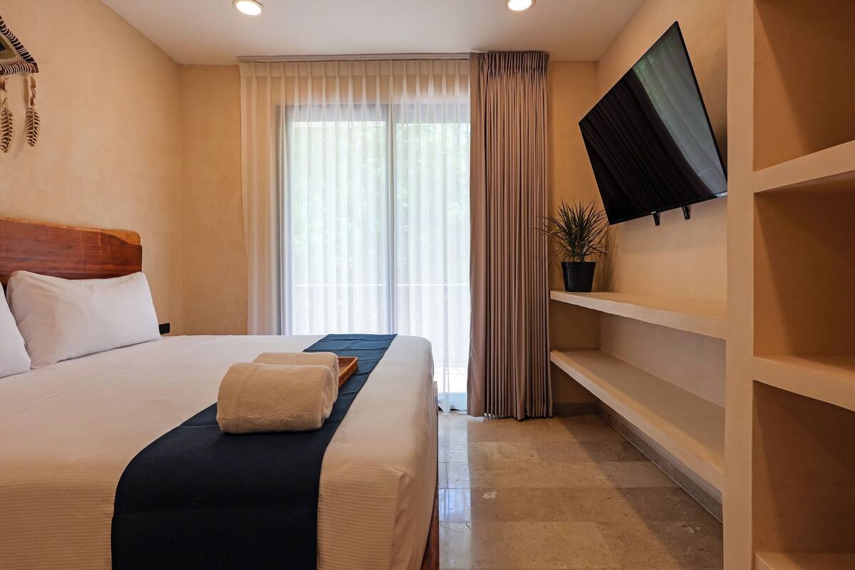 A hotel room with a large bed, a flat-screen TV mounted on the wall, a potted plant, and a window with sheer curtains.