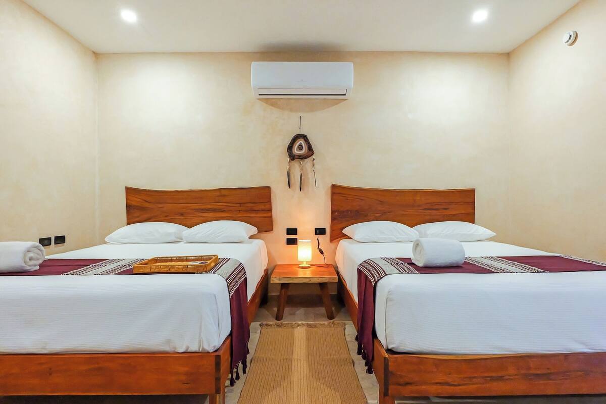 A room with two wooden beds, white bedding, a wall-mounted air conditioner, a small table with a lamp, and a decorative wall hanging.