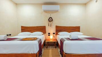 A room with two wooden beds, white bedding, a wall-mounted air conditioner, a small table with a lamp, and a decorative wall hanging.