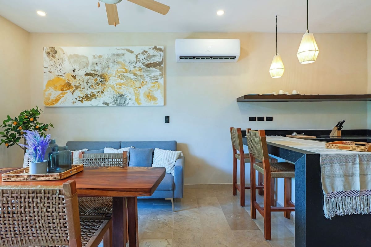 A modern living room with a sofa, dining table, and chairs, a kitchen area with a counter and hanging lights, and a large abstract painting on the wall.