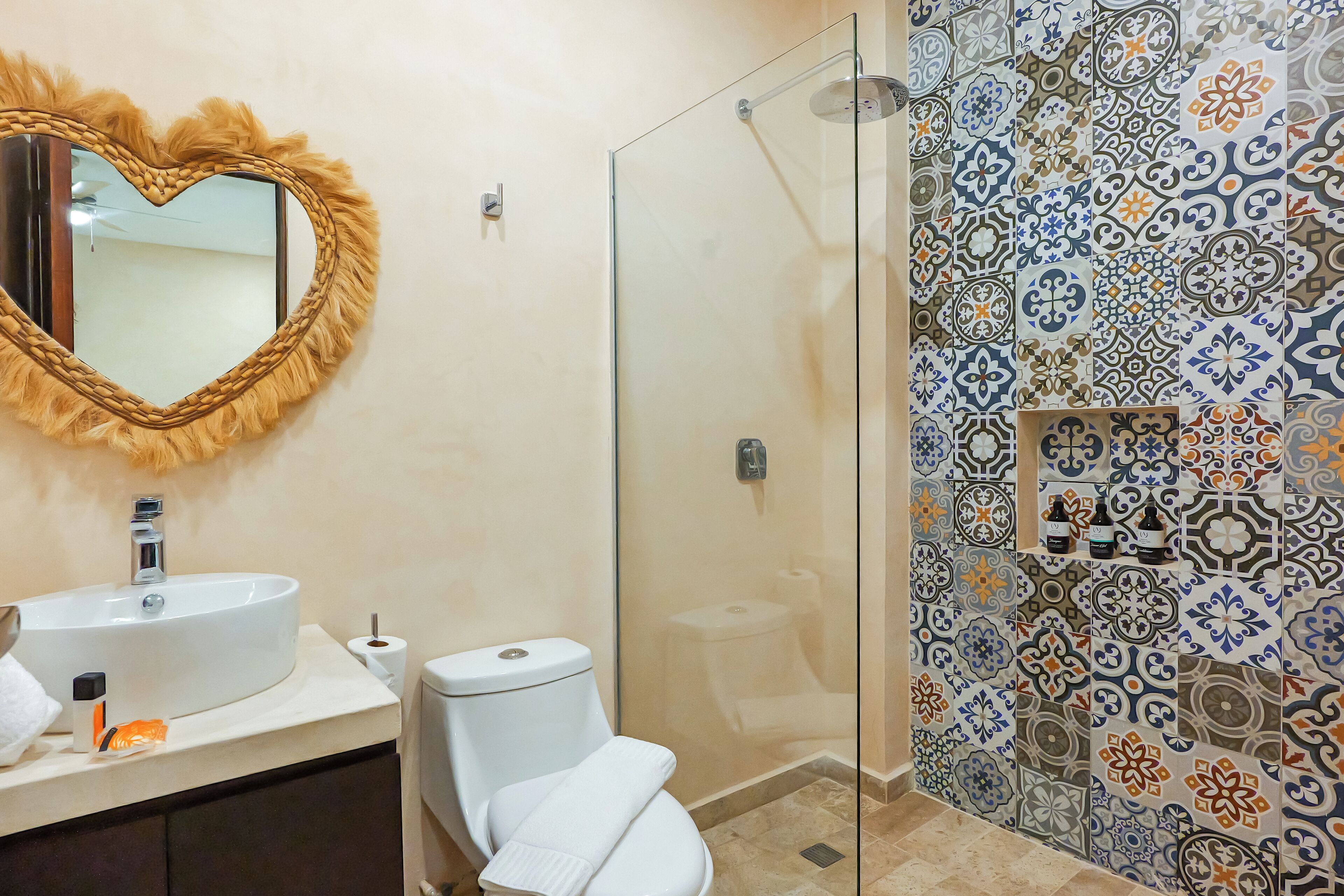 A bathroom with a heart-shaped mirror, a white sink, a toilet, and a shower with a glass door.
