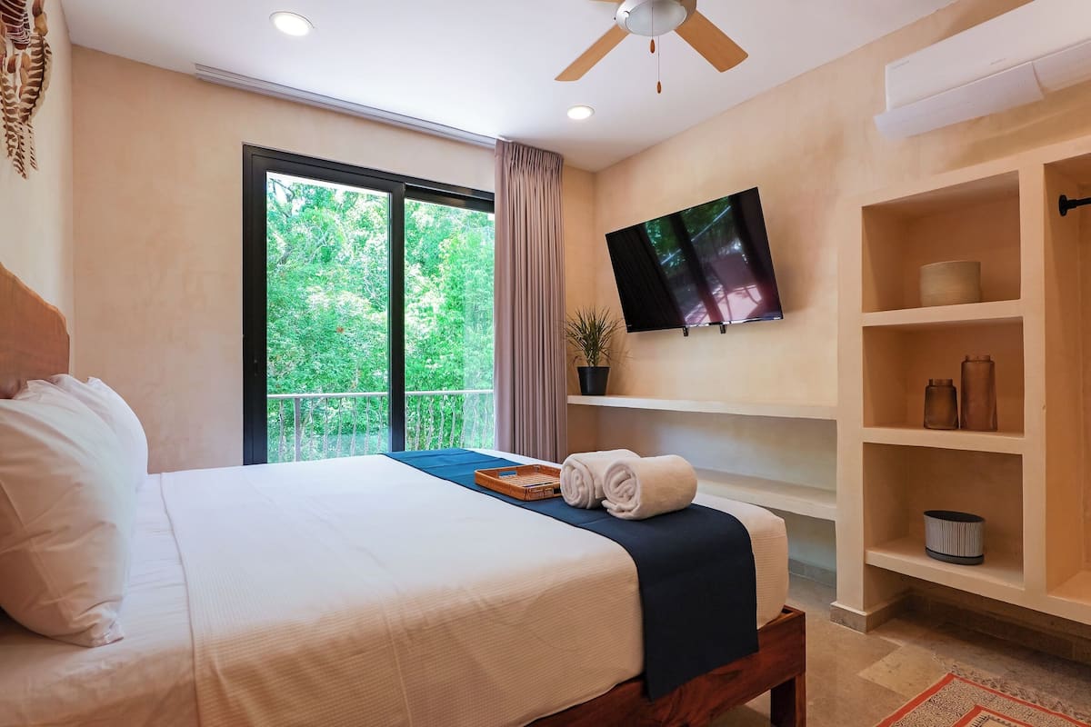 A hotel room with a large bed, a TV mounted on the wall, a balcony with a view of greenery, and a shelf with decorative items.
