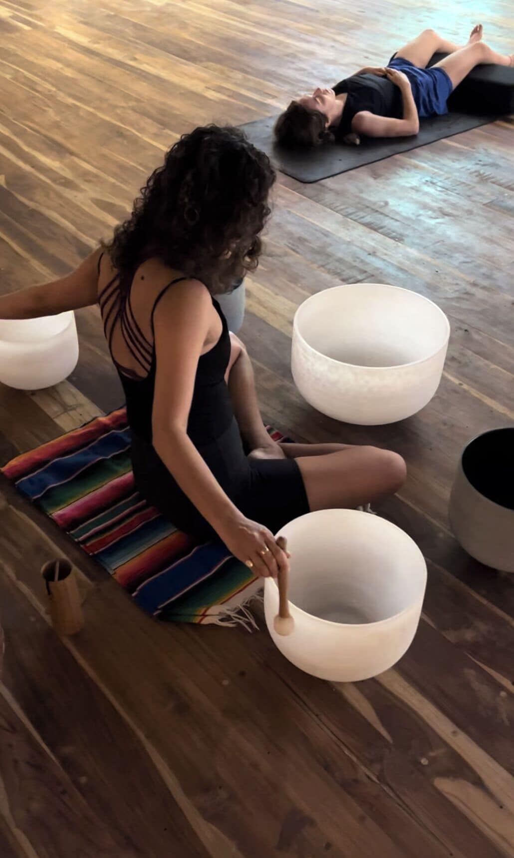 A person sitting on a colorful striped mat, holding a wooden spoon, with large white bowls on the floor.