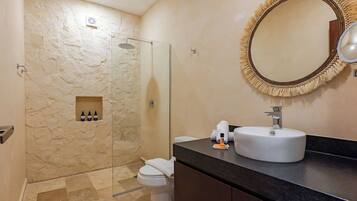 A modern bathroom with a stone-textured wall, a glass-enclosed shower, a white toilet, a black countertop with a sink, and a round mirror with a fringe.