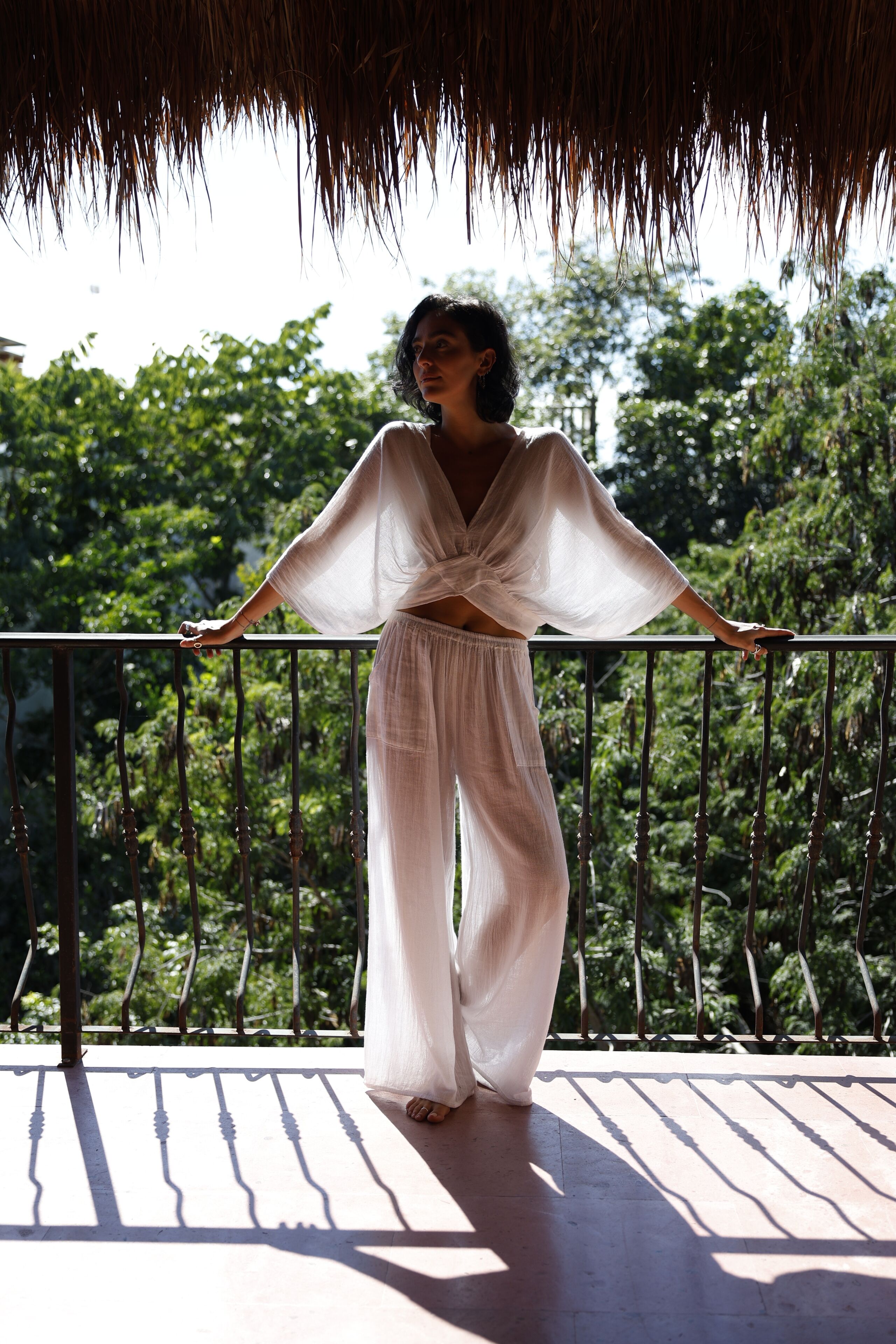 A person standing on a balcony with a railing, trees in the background, and a thatched roof overhead.