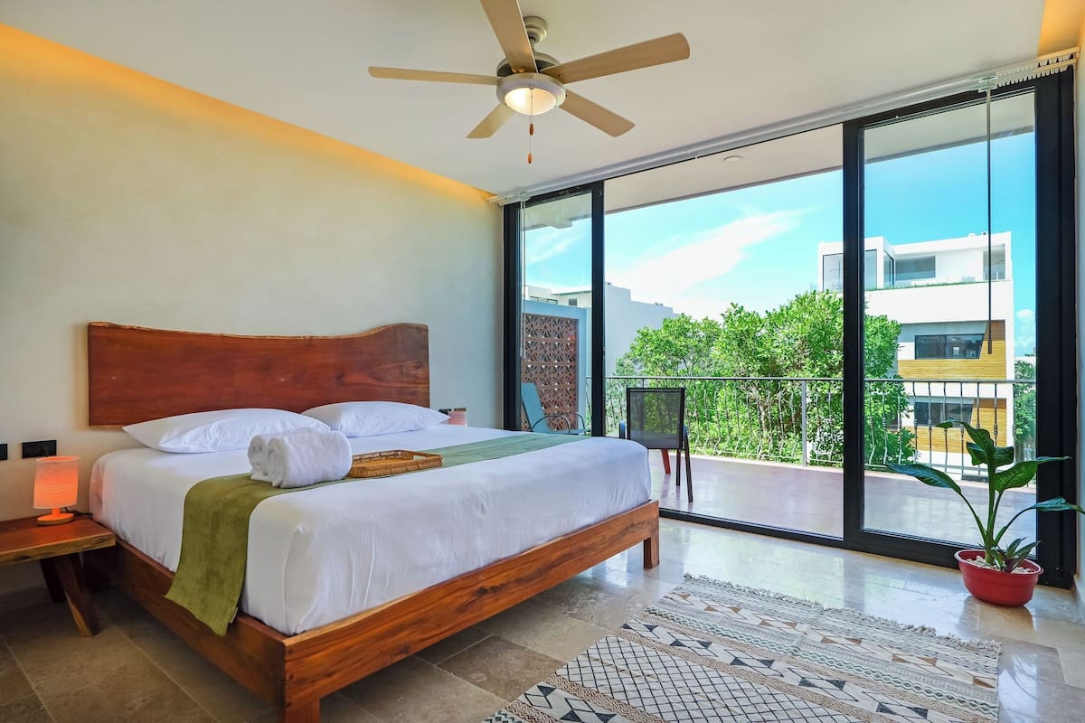 A modern bedroom with a large bed, a ceiling fan, a nightstand, a potted plant, and a balcony with a view of buildings and greenery.