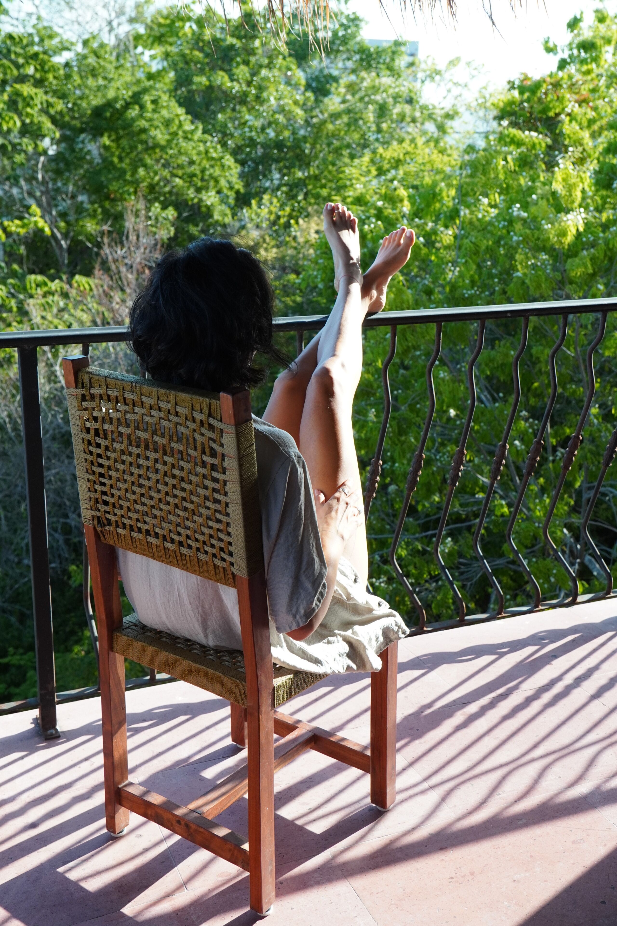 A person seated on a wicker chair with a metal railing, outdoors with greenery in the background.