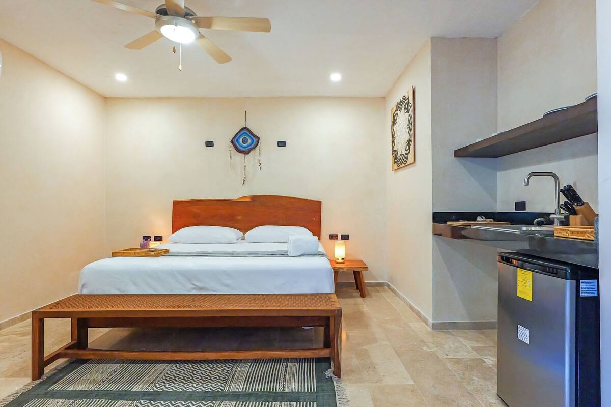 A bedroom with a bed, bedside tables, a bench, a ceiling fan, a wall-mounted artwork, and a kitchenette with a refrigerator and sink.