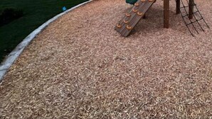 Un parque infantil con tobogán y estructura de escalada, rodeado de un área con acolchado y edificios residenciales al fondo.
