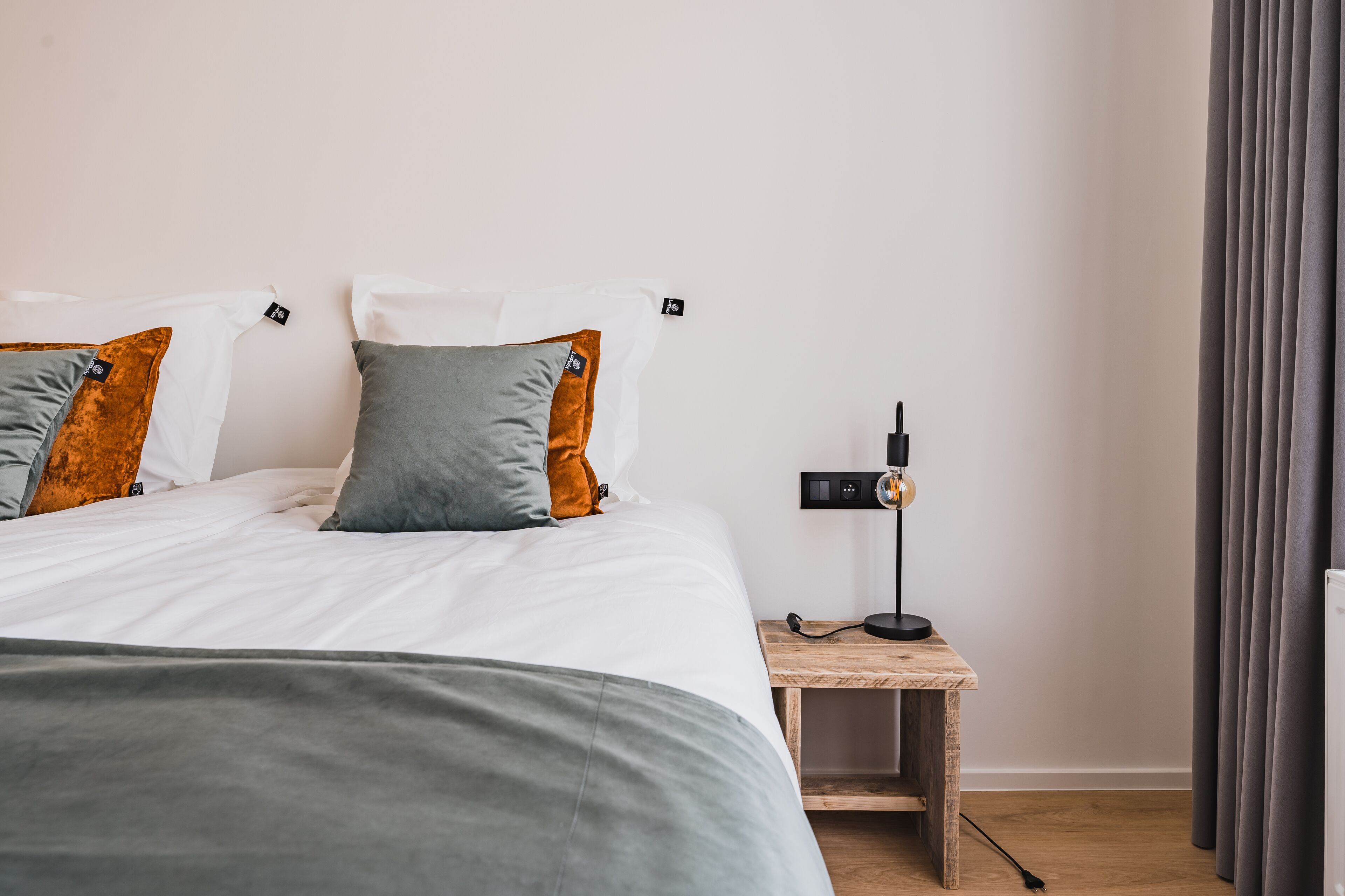 Ein ordentlich bezogenes Bett mit weißer und grauer Bettwäsche, zwei Kissen, ein kleiner Nachttisch mit Lampe und ein grauer Vorhang.
