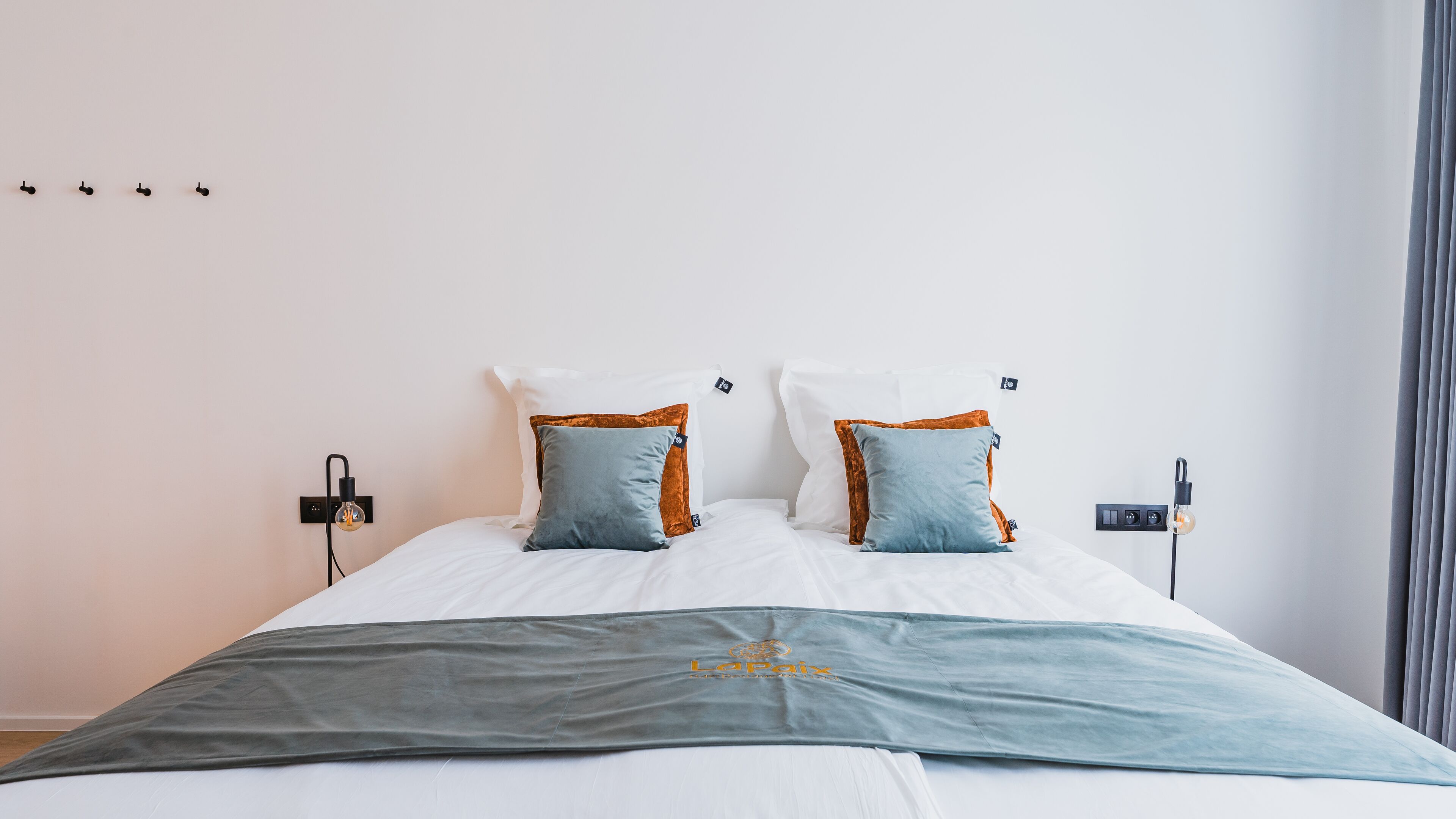 Una cama bien hecha con edredón gris y blanco, dos almohadas con detalles naranja y azul, y una mesita de noche con una lámpara.