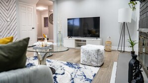 A modern living room with a flat-screen TV, a glass coffee table, a marble ottoman, and a large floor lamp.