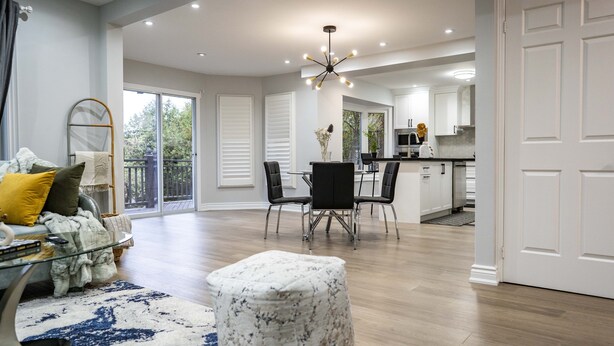 A spacious living area with a glass coffee table, a white ottoman, and a dining table with chairs. A kitchen is visible in the background with white cabinets and a modern design.
