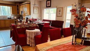 A spacious living area with a dining table set for four, surrounded by red upholstered chairs. A kitchen is visible in the background with wooden cabinets and a window.
