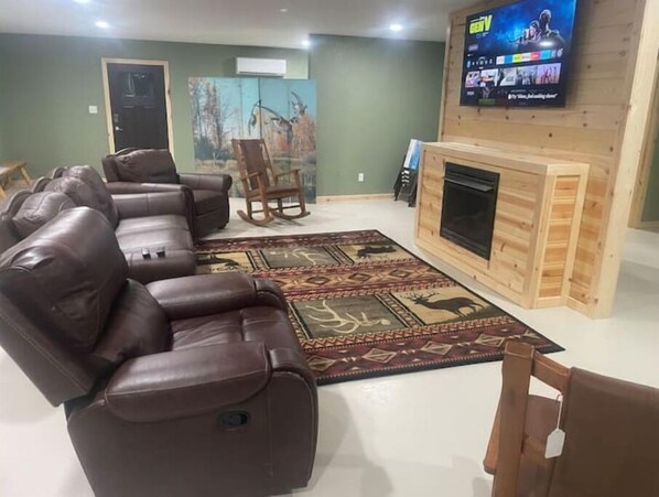 A living room with a brown leather sofa, a wooden fireplace, a TV mounted on the wall, and a painting of deer on the wall. - Puddle Duck Lodge (Corning)