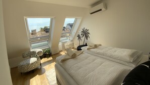 Ein modernes Schlafzimmer mit einem großen Bett, zwei Fenstern, einer Deckenleuchte und Blick auf die Stadt.
