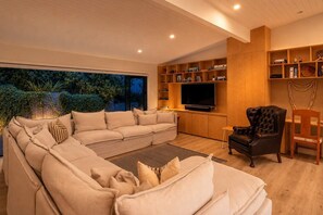 A modern living room with a sectional sofa, a black leather armchair, a television, and wooden shelves with various items. - Afio Mai - Walk to Little Oneroa Beach (Waiheke Island)
