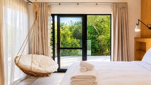 Habitación de hotel con una cama, una silla colgante, una mesita de noche de madera y vista a la vegetación por la ventana.