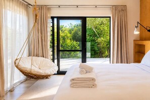 Hotellihuone, jossa on sänky, riipputuoli, puinen yöpöytä ja ikkunasta avautuva näkymä vehreään maisemaan.