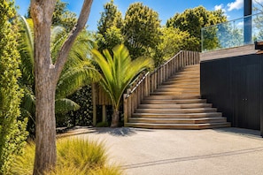 Un escalier menant à un bâtiment doté d’une rambarde en verre, le tout entouré de verdure.