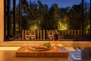 A wooden table with a bottle of Man O' War wine, two glasses of wine, and a plate of grapes and crackers by a window with a view of trees. - Afio Mai - Walk to Little Oneroa Beach (Waiheke Island)
