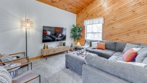 Ein modernes Wohnzimmer mit grauem Sofa, Holzboden, Flachbildfernseher, Stehlampe und einem Fenster mit Vorhängen.