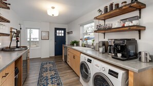 Een kleine keuken met houten kastjes, een wasmachine, een koffiezetapparaat en planken met potten.
