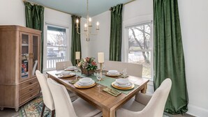 Een eetkamer met een houten tafel gedekt voor vier personen, groene gordijnen, een kroonluchter en een kast met glazen deuren.
