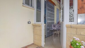 Un estrecho pasillo exterior con suelo de baldosas, una pequeña mesa con sillas y una planta en maceta.