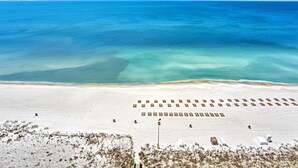 Eine Vogelperspektive zeigt einen Strand mit Liegestühlen und Sonnenschirmen, feinen Sand und einem klaren, blauen Meer.