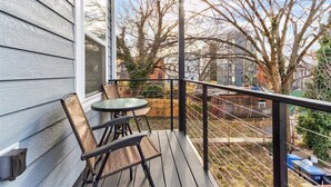 Un balcón con una mesa y dos sillas, con vistas a un patio cercado con árboles y un edificio al fondo.