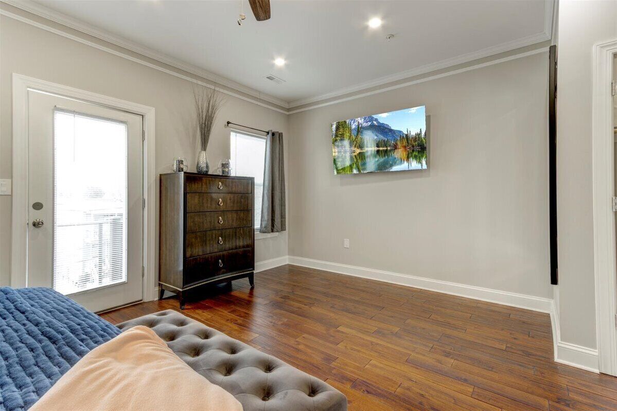 A spacious room with hardwood flooring, a large bed with a tufted headboard, a dark wooden dresser, and a wall-mounted painting of a mountain lake.