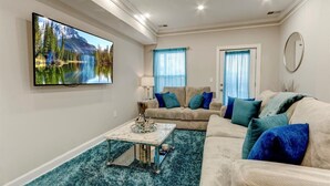 Una sala de estar con un televisor de pantalla plana grande, un sofá color beige, una mesa de centro y una alfombra azul.