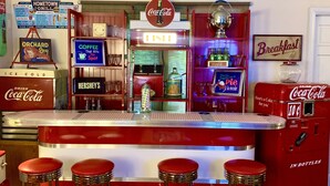 Un bar estilo retro con una máquina expendedora de Coca-Cola, un jukebox y un cartel que anuncia 'Breakfast'.