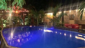 Una piscina exterior con una fuente, rodeada de palmeras y una zona cubierta con luces colgantes.