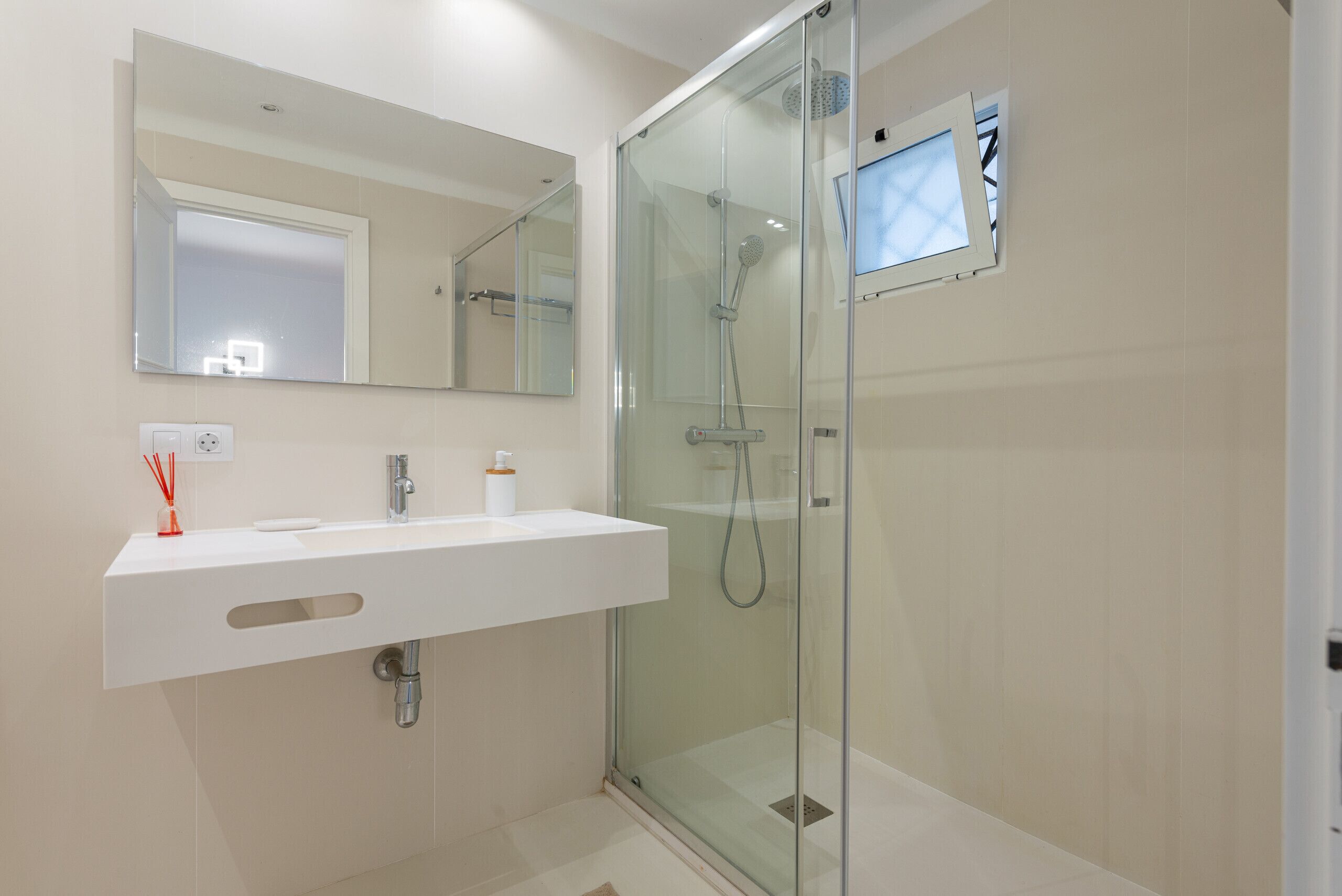A modern bathroom with a glass-enclosed shower, a white sink with a mirror above, and a small window.