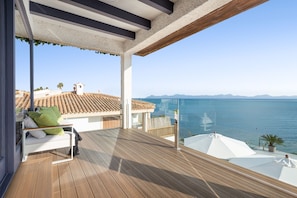 Eine Holzterrasse mit einem weißen Sofa und grünen Kissen, mit Blick auf das klare, blaue Meer und ferne Berge.
