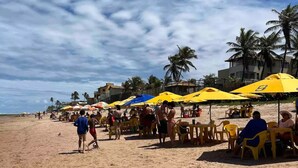 A beach with yellow umbrellas and tables, palm trees, and buildings in the background. - Cobertura C/suíte - Salvador à 2km Praia Aeroporto (Centro Lauro de Freitas)