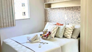 A neatly made bed with a white comforter and pillows, a bedside table with a phone and a basket of snacks, and a window with blinds. - Cobertura C/suíte - Salvador à 2km Praia Aeroporto (Centro Lauro de Freitas)