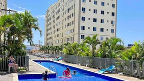 A swimming pool with a yellow float, surrounded by palm trees and a tall building in the background. - Cobertura C/suíte - Salvador à 2km Praia Aeroporto (Centro Lauro de Freitas)