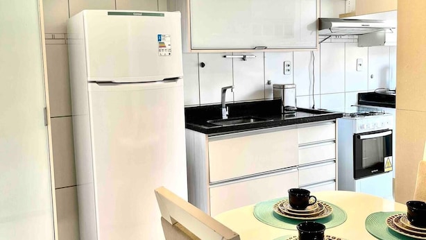 A modern kitchen with white cabinets, a built-in oven, and a refrigerator. There is a dining table with placemats and cups. - Cobertura C/suíte - Salvador à 2km Praia Aeroporto (Centro Lauro de Freitas)
