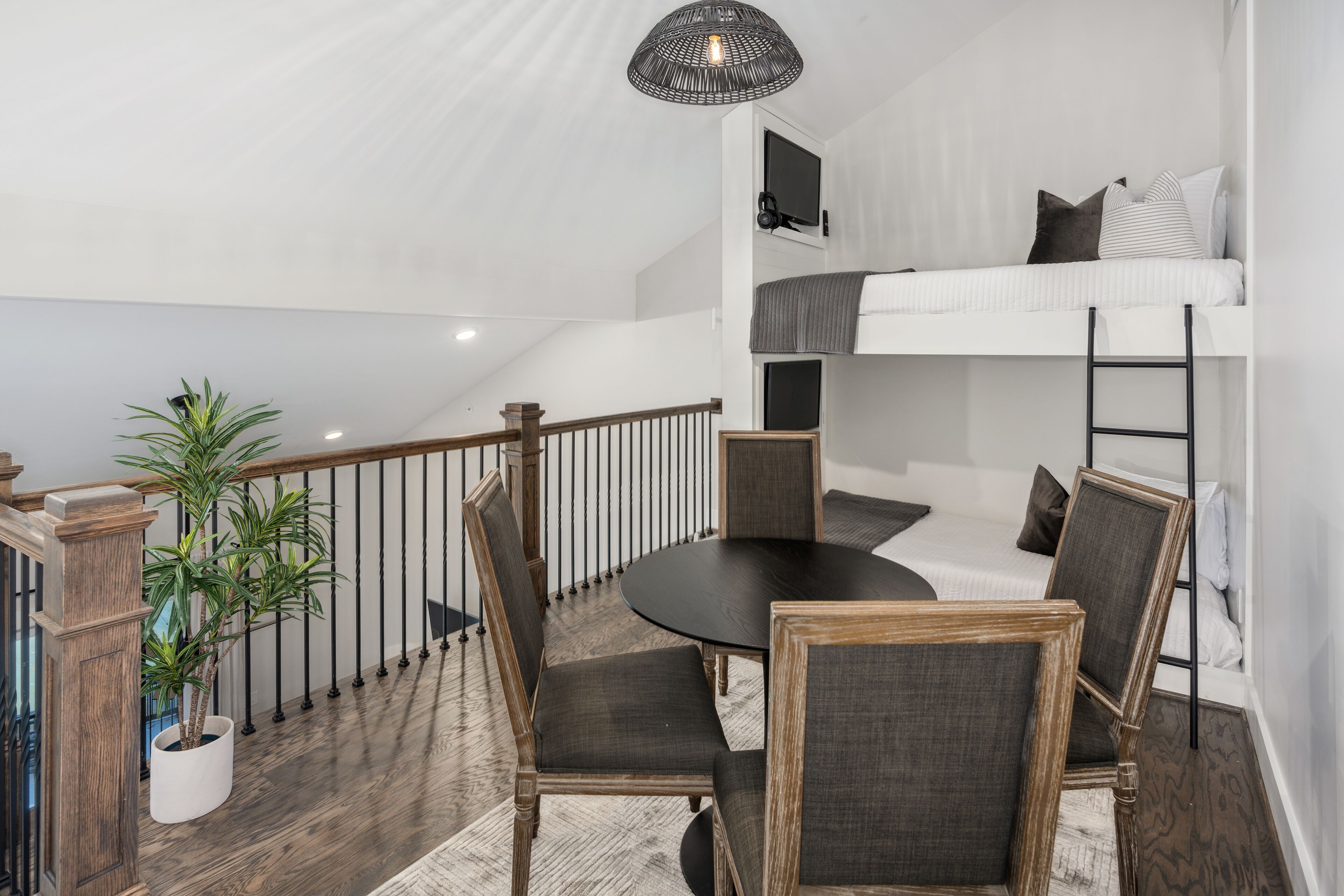 A modern loft bedroom with a bunk bed, a dining table with chairs, a potted plant, and a staircase.