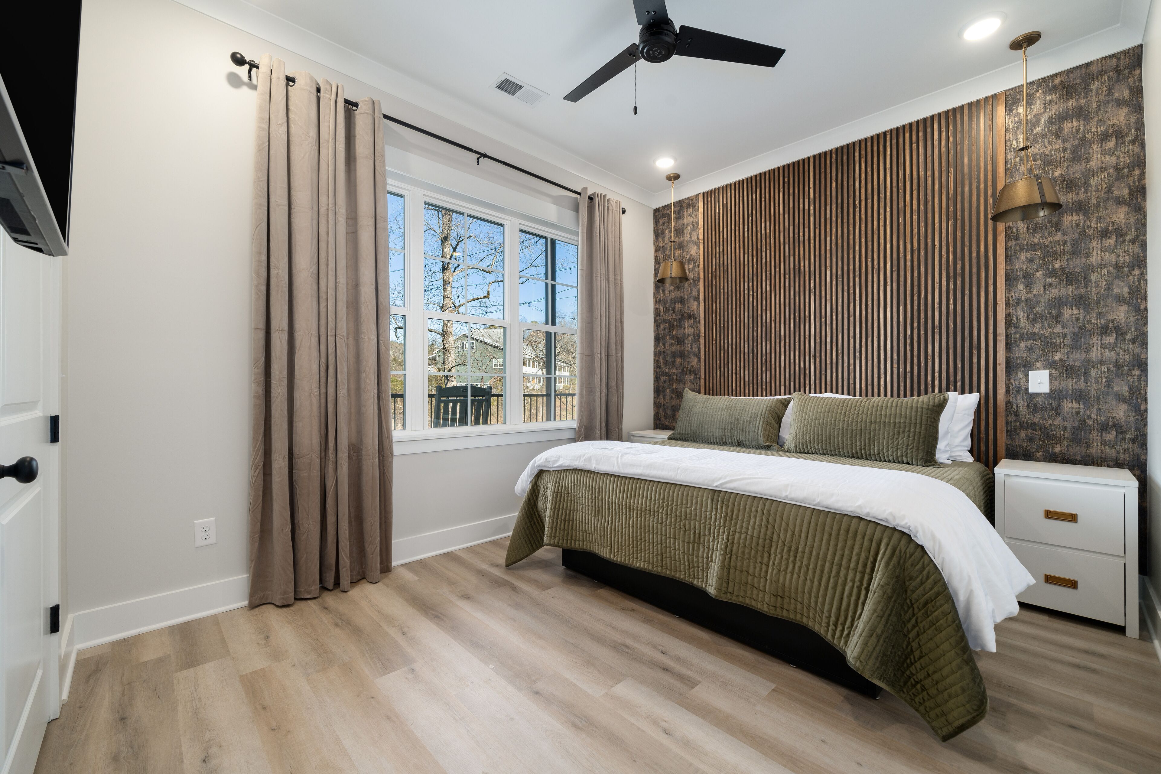 A bedroom with a large bed, a ceiling fan, a window with curtains, and a view of the outside.